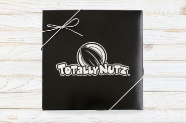 black square box with silver totally nutz logo embossed on top and closed with a silver ribbon, box has gingham print bottom and the photo is taken on a white wood background 